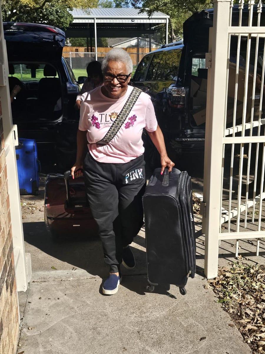 A smiling Dallas Chapter Lady in a pink TLOD shirt wheeling suitcases through a gate, with vehicles loaded with boxes behind her