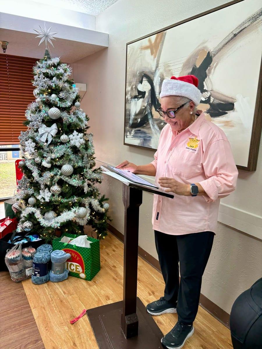 A Dallas Chapter Lady in a pink chapter shirt and Santa hat reading at a lectern beside a decorated holiday tree and gifts of blankets
