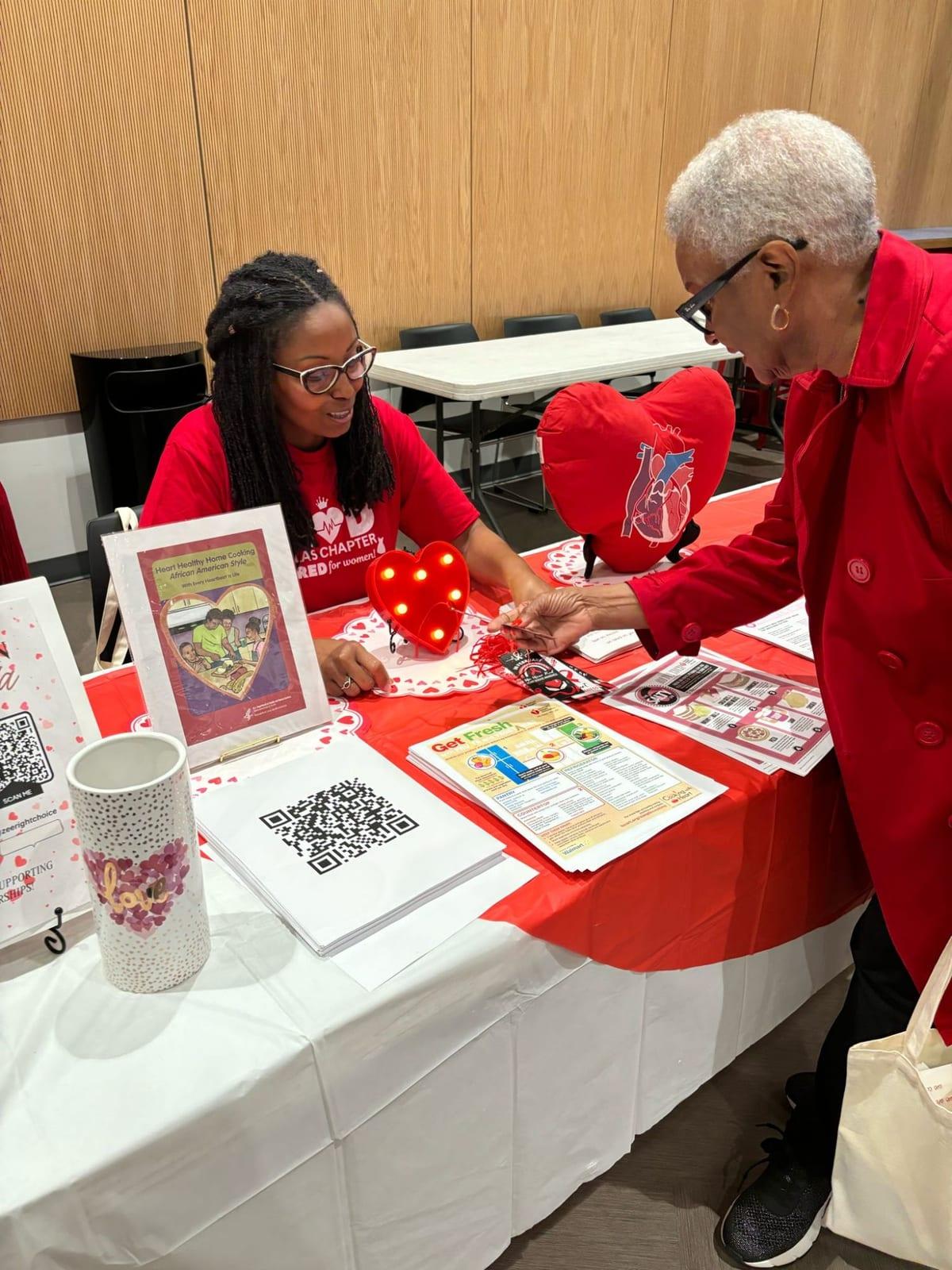 A Dallas Chapter Lady in a Go Red for Women shirt sharing heart health materials with a woman in a red coat at a table decorated with hearts