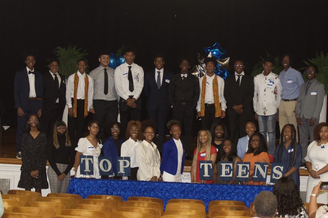 Dallas Top Teens in formal dress gathered on stage behind a table spelling TOP TEENS, with blue and silver balloons